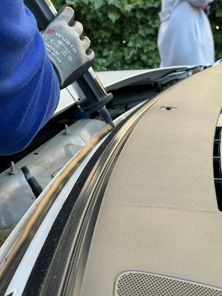 Close-up of a technician applying sealant to a car windshield in daylight.