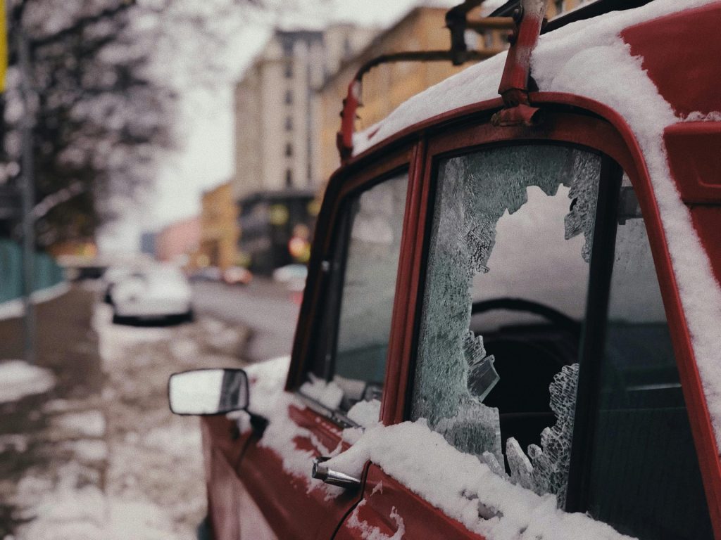 A red car with a broken window parked in a snowy urban street, highlighting winter weather impact.